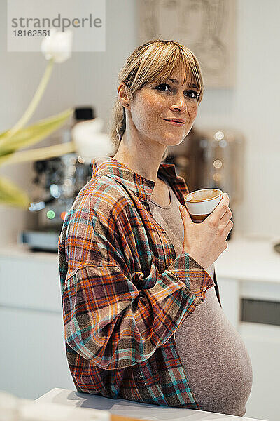 Nachdenkliche schwangere Frau trägt kariertes Hemd und hält zu Hause eine Tasse in der Hand