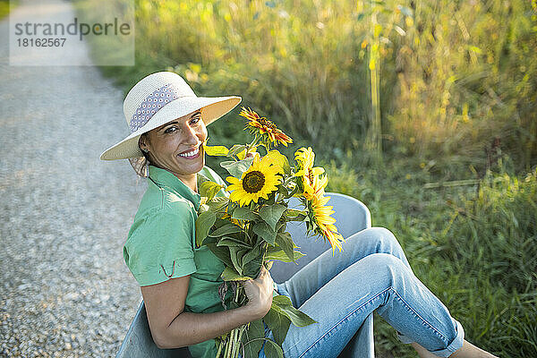 Glücklicher reifer Gärtner mit einem Strauß Sonnenblumen  der in einer Schubkarre sitzt