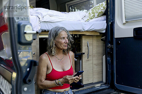 Lächelnde ältere Frau sitzt mit Smartphone auf der Stoßstange eines Wohnmobils