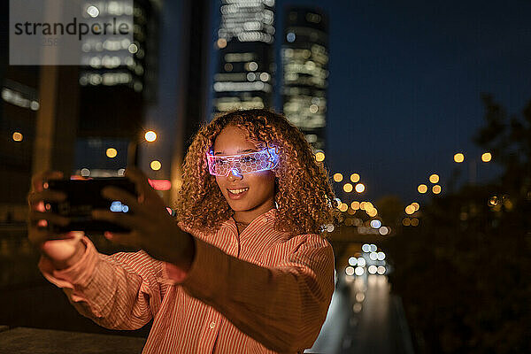 Lächelnde Frau mit leuchtender Smart-Brille  die nachts ein Selfie mit dem Smartphone macht