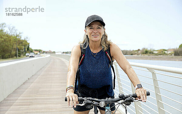 Glückliche ältere Frau mit Mütze  die auf der Promenade Fahrrad fährt