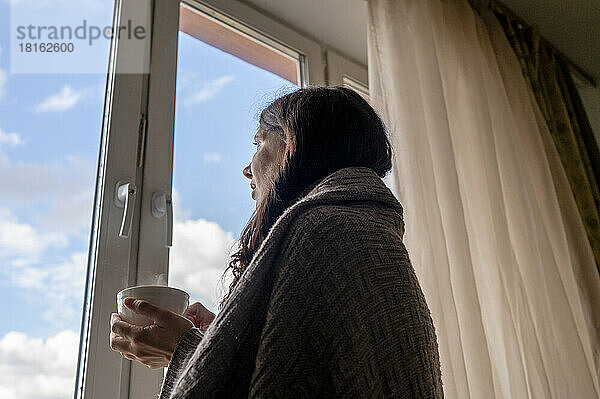 Frau in Decke gehüllt  hält Kaffeetasse in der Hand und blickt zu Hause durchs Fenster