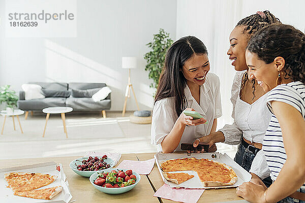 Fröhliche Mitbewohner essen Pizza und nutzen zu Hause ihr Smartphone