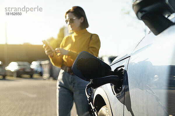 Elektroauto wird an der Station aufgeladen  während eine Frau an einem sonnigen Tag ihr Smartphone im Hintergrund benutzt