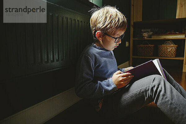 Junge mit Brille sitzt zu Hause auf dem Boden und liest ein Buch