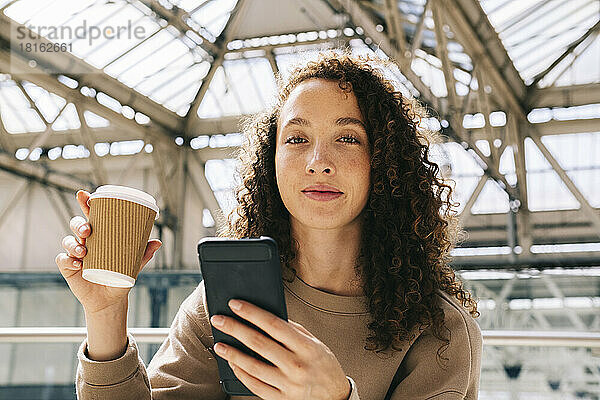 Lächelnde Frau sitzt mit Handy und trinkt Kaffee am Bahnhof