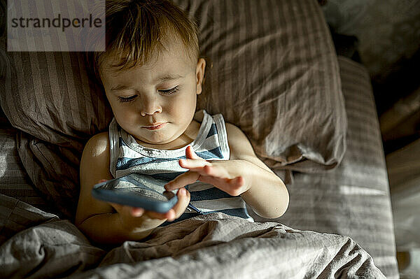 Niedliches Kleinkind benutzt Smartphone im Bett im Schlafzimmer