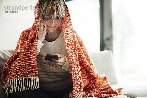 Lächelnde Frau mit Decke und Smartphone zu Hause