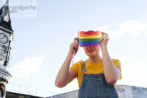 Lächelnde nicht-binäre Person  die ihr Gesicht mit einer Regenbogenfahne bedeckt