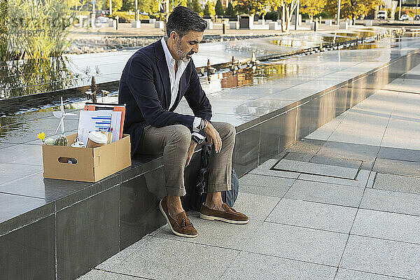 Trauriger Geschäftsmann mit Büromaterial in einer Kiste  die an der Wand sitzt
