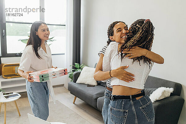 Frau mit Pizzakarton blickt auf Mitbewohner  die sich im Wohnzimmer umarmen