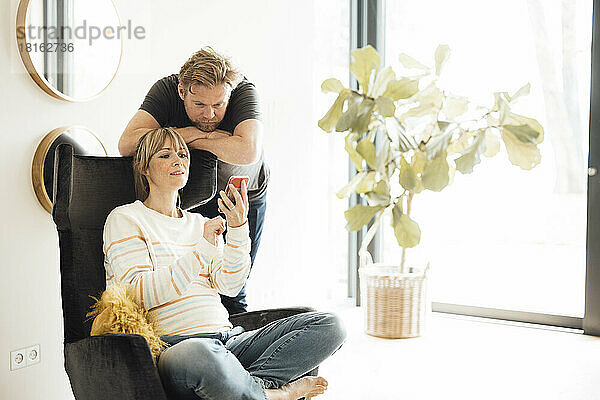 Schwangere Frau sitzt auf Stuhl und teilt Smartphone mit Mann zu Hause