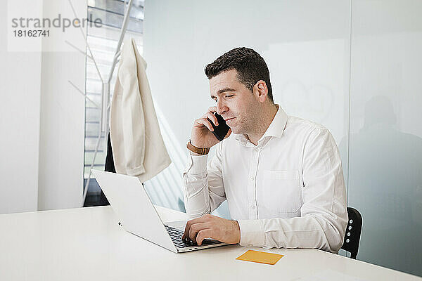 Junger Geschäftsmann spricht am Schreibtisch im Büro mit Smartphone und Laptop