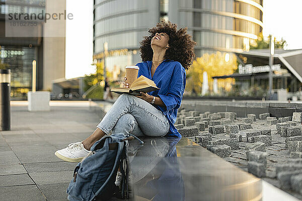Fröhliche Afro-Frau mit Buch und Einwegbecher sitzt an der Wand