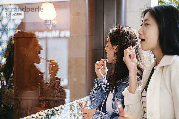 Lesbian friends applying lip gloss looking in glass wall