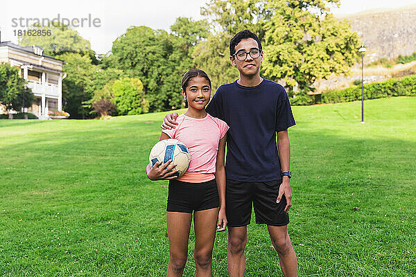 Junge mit Arm um Schwester  die Ball hält  der auf Gras im Rasen steht