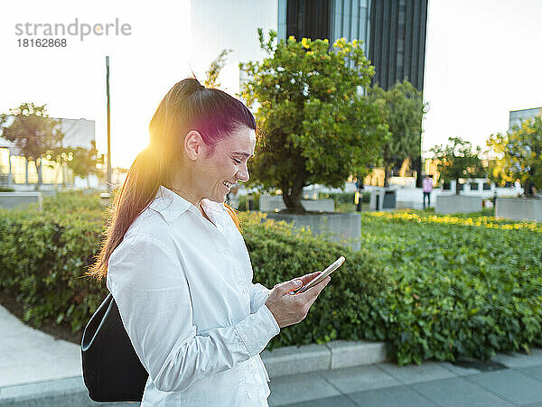 Glückliche Geschäftsfrau nutzt Smartphone im Büropark bei Sonnenuntergang