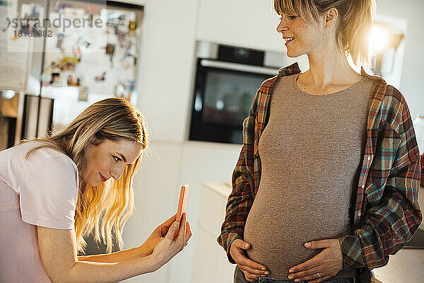 Lächelnde Schwester fotografiert zu Hause mit dem Handy den Bauch einer schwangeren Frau