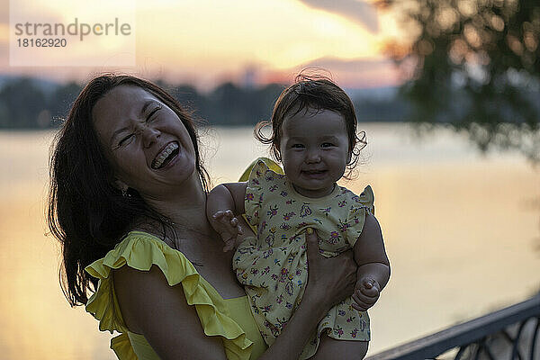 Glückliche Mutter trägt ihre Tochter bei Sonnenuntergang vor dem Fluss