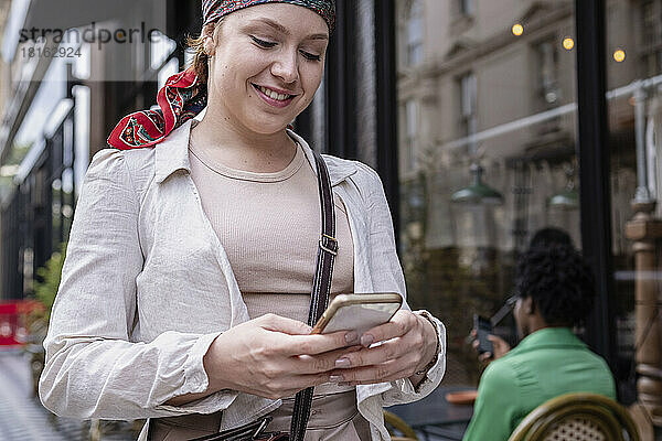 Glückliche junge Frau mit Bandana und Smartphone im Straßencafé