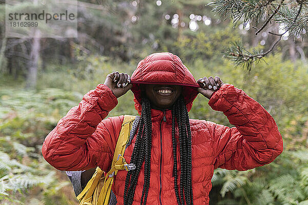 Glückliche Frau  die ihr Gesicht mit der Kapuze einer Jacke bedeckt und im Wald steht