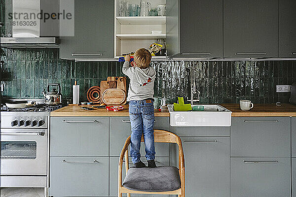 Boy searching in kitchen cabinet standing on chair at home