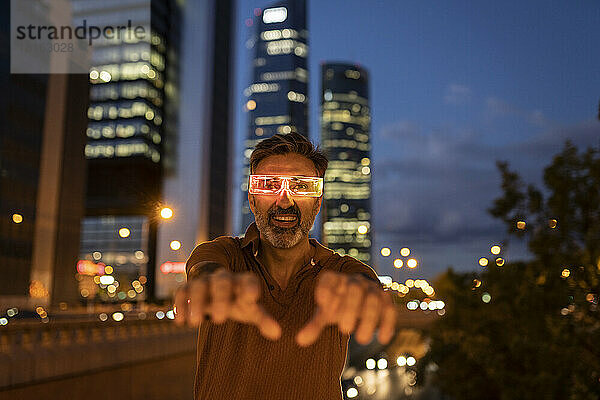 Glücklicher Geschäftsmann gestikuliert mit futuristischer Brille nachts in der Stadt