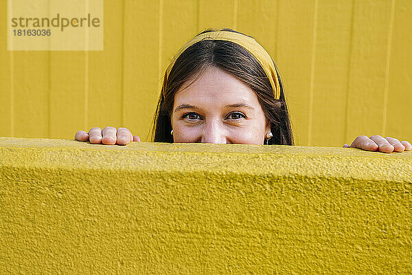 Happy woman looking from behind yellow wall