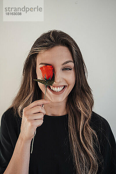 Glückliche junge Frau bedeckt ihr Auge mit einer Rose vor weißem Hintergrund