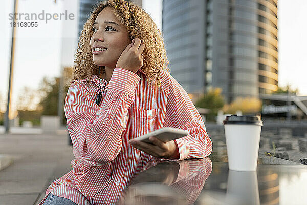 Lächelnde Frau mit Smartphone und In-Ear-Kopfhörern neben Bank