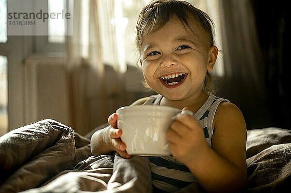 Glücklicher kleiner Junge mit Tasse im Bett zu Hause