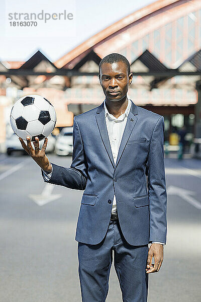 Junger Geschäftsmann mit Fußball steht an sonnigem Tag auf dem Parkplatz