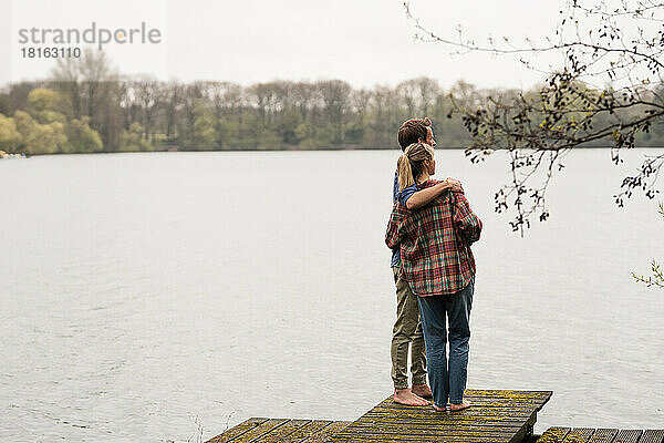 Mann mit Arm um Frau  die auf einem Steg steht und auf den See blickt