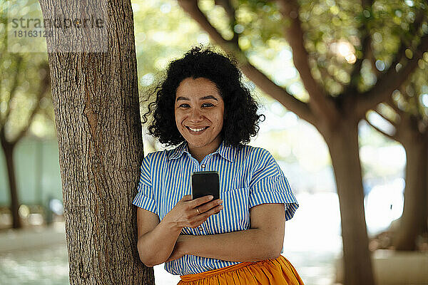 Glückliche Frau mit Mobiltelefon lehnt an Baumstamm
