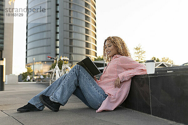 Lächelnde Frau liest ein Buch und sitzt auf einem Fußweg in der Stadt
