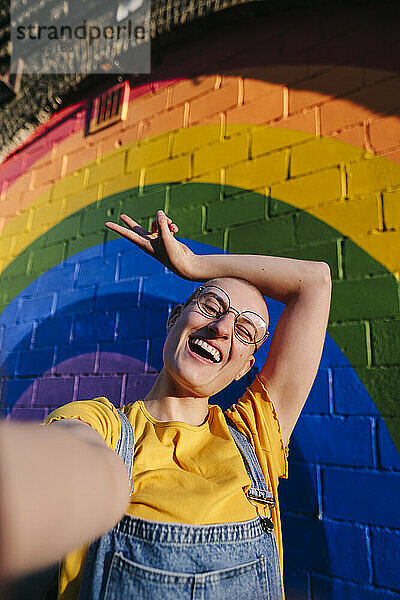 Fröhliche Transgender-Person gestikuliert mit einem Friedenszeichen und macht ein Selfie vor der Regenbogenwand