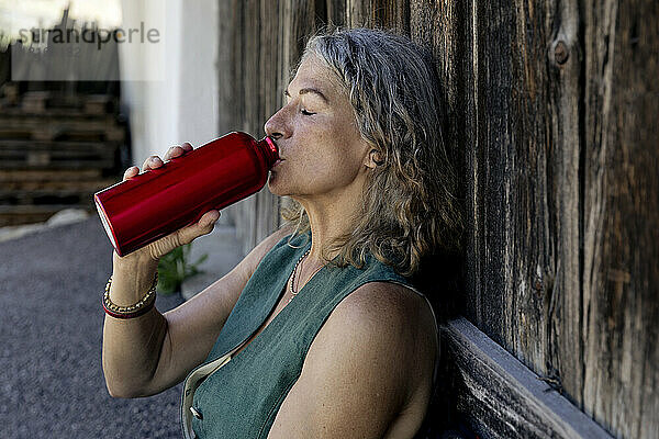 Ältere Frau mit geschlossenen Augen trinkt Wasser aus der Flasche an der Holztür