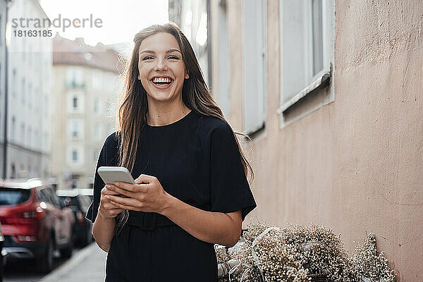 Fröhliche junge Frau mit Smartphone steht an der Wand