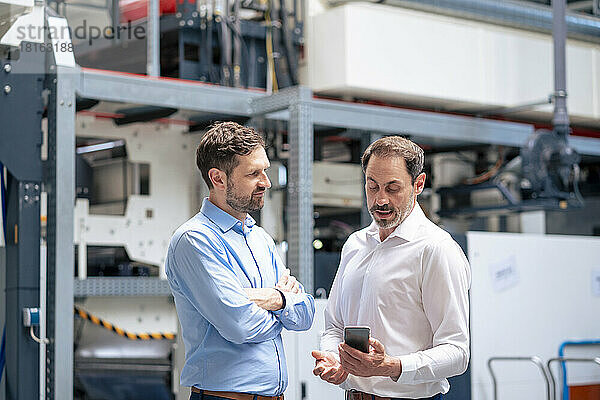 Geschäftsmann schaut auf sein Smartphone und diskutiert mit Kollegen in der Fabrik