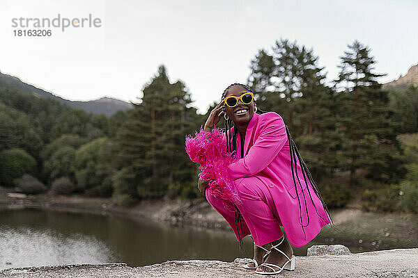 Glückliche junge Frau mit Sonnenbrille kauert vor dem See