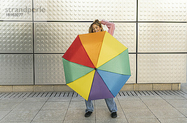 Young woman with hand in hair holding colorful umbrella on footpath