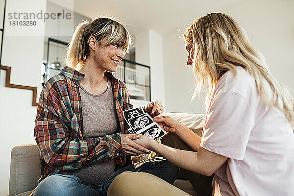 Excited pregnant woman showing ultrasound to sister at home