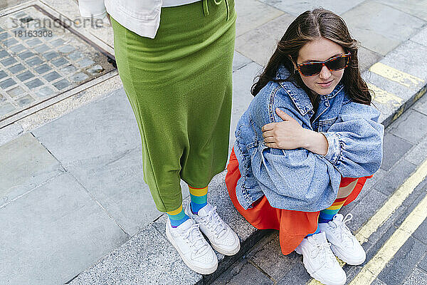 Woman standing by lesbian friend wearing sunglasses sitting on footpath