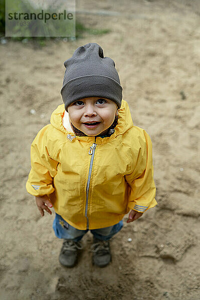 Niedlicher kleiner Junge mit Strickmütze und gelber Regenjacke auf Sand