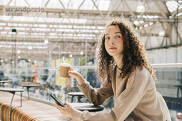 Junge Frau sitzt mit Handy und trinkt Kaffee am Bahnhof