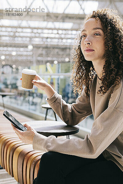 Nachdenkliche Frau sitzt mit Handy und trinkt Kaffee am Bahnhof