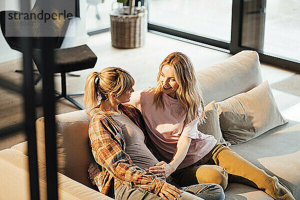 Lächelnde Frau berührt den Bauch einer schwangeren Frau  die zu Hause auf dem Sofa sitzt