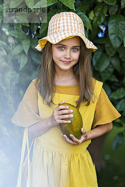 Lächelndes Mädchen mit Hut steht mit Mango vor der Pflanze