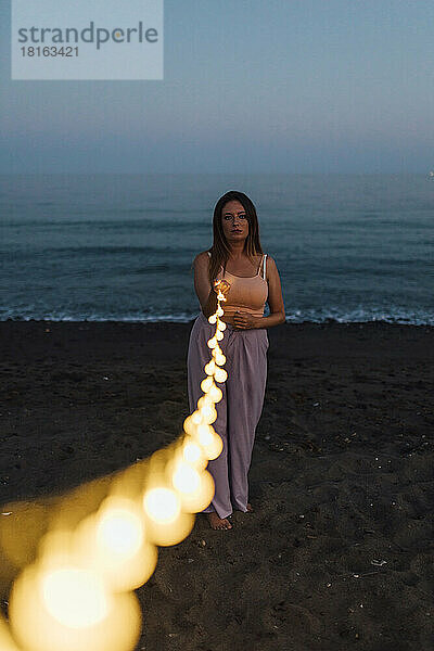Frau hält beleuchtete Lichterkette am Strand