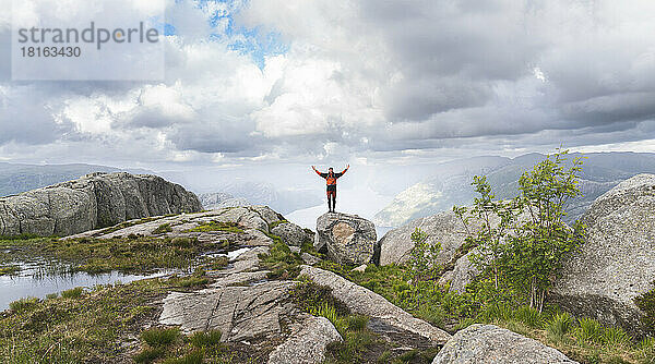 Junger Wanderer mit erhobenen Armen steht auf einem Felsen unter bewölktem Himmel